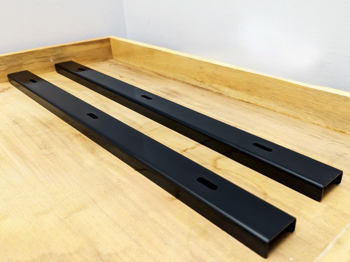 A pair of black slotted steel C-channel table strengtheners on an oak tabletop