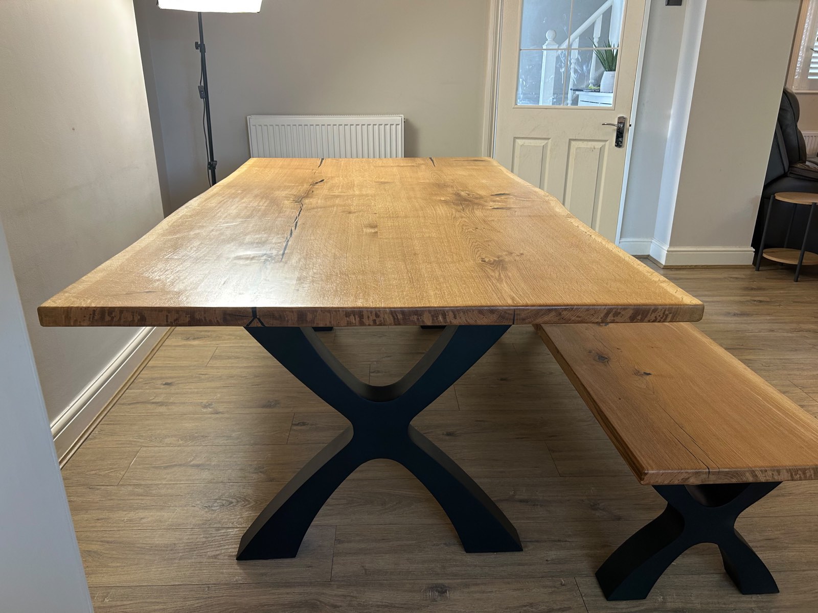 French oak dining table photographed end on, both black Designer X legs visible with the bench alongside